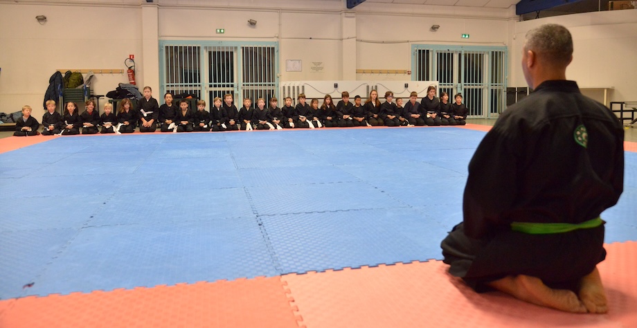 Une trentaine d'enfants assis en ligne sur leurs pieds, face à l'enseignant